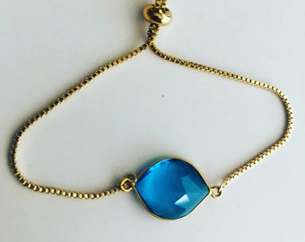 May include: A gold chain bracelet with a blue gemstone pendant. The bracelet has an adjustable sliding clasp.