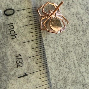 Op de afbeelding: Een ros&eacute;goudkleurige oorsteker in de vorm van een spin met een wit parelachtig lichaam, geplaatst naast een liniaal die een breedte van ongeveer 1,3 cm aangeeft. De spin heeft acht dunne poten en een centrale stift voor bevestiging.