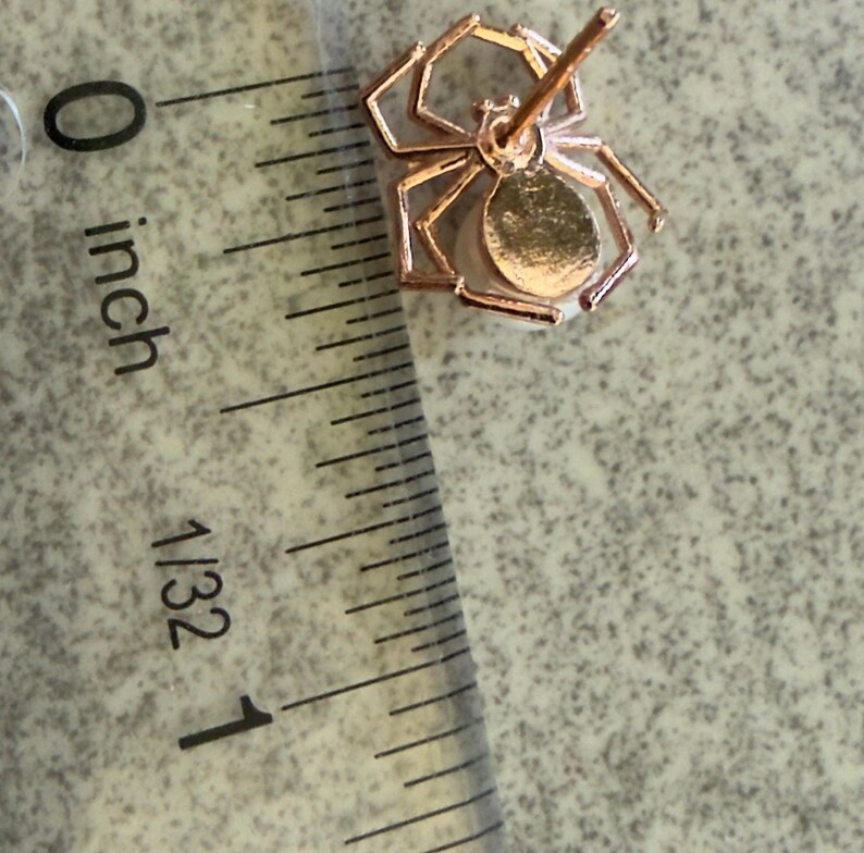 Op de afbeelding: Een ros&eacute;goudkleurige oorsteker in de vorm van een spin met een wit parelachtig lichaam, geplaatst naast een liniaal die een breedte van ongeveer 1,3 cm aangeeft. De spin heeft acht dunne poten en een centrale stift voor bevestiging.