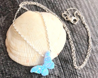 May include: A silver chain necklace with a blue opal butterfly pendant. The necklace is displayed on a seashell.