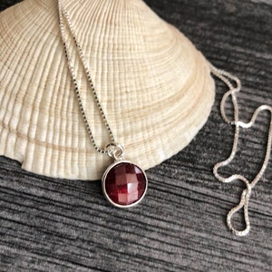 May include: A silver chain necklace with a red gemstone pendant. The pendant is a round, faceted stone set in a silver bezel.