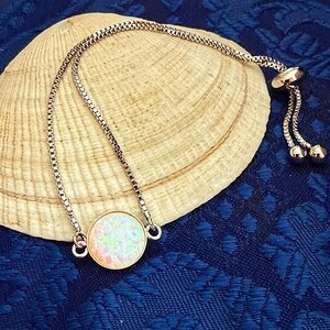 May include: A rose gold tone adjustable bracelet featuring a circular pendant with a white iridescent opal-like stone. The bracelet has a delicate box chain design with two small round beads at the ends of the sliding closure mechanism.