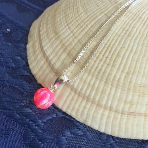 May include: A silver necklace with a small, round, pink opal pendant. The pendant is a vibrant shade of pink and hangs from a delicate silver chain. The necklace is displayed on a large, textured seashell against a blue background.