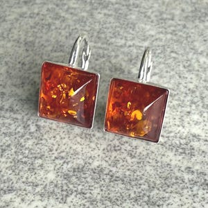 May include: A pair of square amber earrings with silver-colored hooks. The amber stones have a rich, warm brown color with golden inclusions. The earrings are set in a silver-toned metal frame.