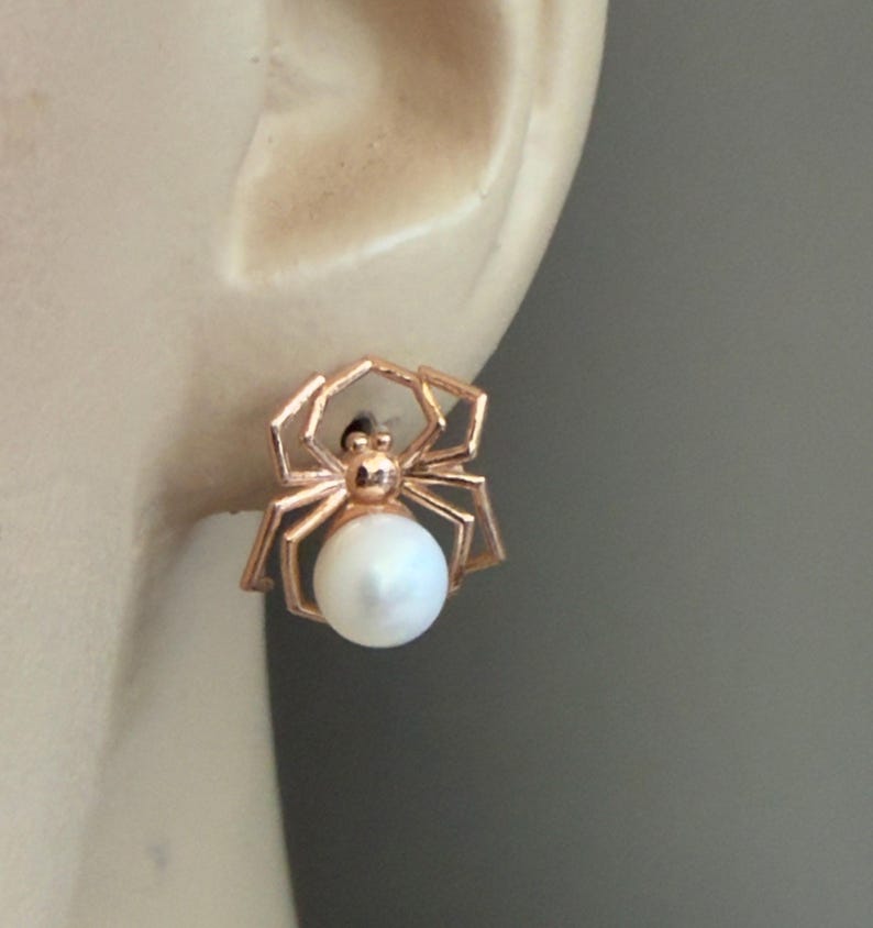 Op de afbeelding: Een ros&eacute;goudkleurige spinnen-oorsteker met een gepolijst metalen frame in de vorm van een spin met hoekige poten en een ronde, witte, glanzende parel die het achterlijf van de spin vormt, bevestigd aan een oorlel met een neutrale kleur.