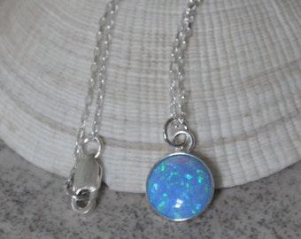 May include: A silver necklace featuring a round, blue opal pendant. The pendant has a shimmering, speckled appearance and is set in a silver bezel. The chain is delicate and has a simple clasp. The necklace is displayed on a neutral background.