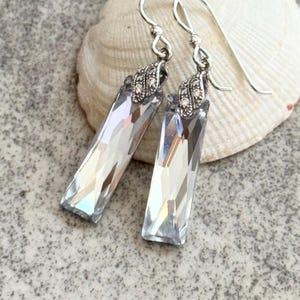 Może przedstawiać: Para prostokątnych kolczyków z kryształkami z haczykami w kolorze srebrnym. Przezroczyste kryształy mają wiele faset, odbijających światło. Górna część każdego kolczyka ma ozdobną srebrną nasadkę z małymi zdobieniami. Kolczyki są prezentowane na muszli.