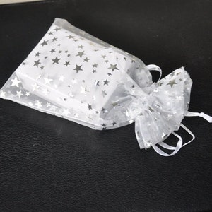 Op de afbeelding: Een rechthoekige witte doos in een doorzichtig wit organza zakje met trekkoord. Het zakje is voorzien van een herhalend patroon van kleine, metallic zilveren sterren op de doorschijnende stof, afgewerkt met witte linten bij de opening.