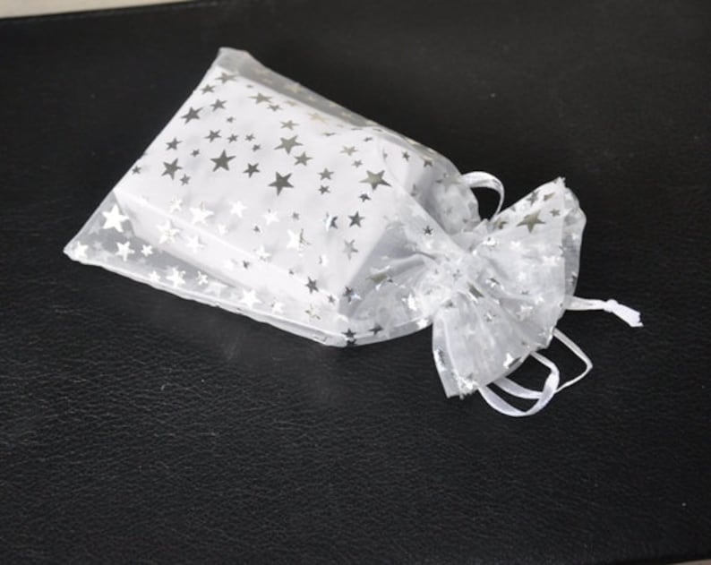 Op de afbeelding: Een rechthoekige witte doos in een doorzichtig wit organza zakje met trekkoord. Het zakje is voorzien van een herhalend patroon van kleine, metallic zilveren sterren op de doorschijnende stof, afgewerkt met witte linten bij de opening.
