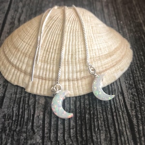 May include: A pair of silver dangle earrings with a crescent moon charm made of white opal. The earrings are hanging from a seashell.