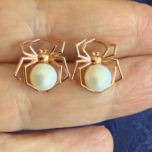 Op de afbeelding: Een paar oorknopjes in de vorm van spinnen, gemaakt van ros&eacute;goudkleurig metaal. Elke spin heeft een ronde, witte, iriserende parel als achterlijf en een kleine, gladde metalen kop met acht hoekige poten die naar buiten toe uitsteken.