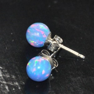 May include: A pair of silver stud earrings with blue opal stones. The opals have a light blue base with iridescent pink, green, and blue flashes.