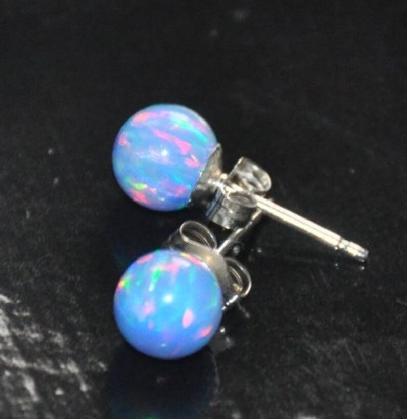 May include: A pair of silver stud earrings with blue opal stones. The opals have a light blue base with iridescent pink, green, and blue flashes.