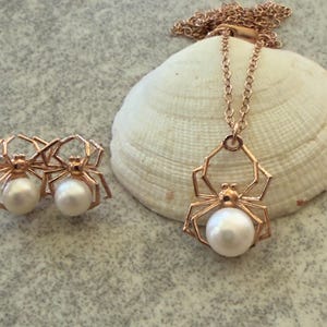 Op de afbeelding: Een sieradenset bestaande uit een ketting en een paar oorknopjes, elk met een ros&eacute;goudkleurig spinnenmotief met een witte parel als lichaam. De ketting heeft een fijne schakelketting en de oorbellen zijn ontworpen als bijpassende spinnenknopjes.