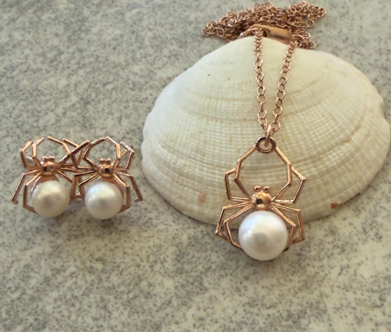 Op de afbeelding: Een sieradenset bestaande uit een ketting en een paar oorknopjes, elk met een ros&eacute;goudkleurig spinnenmotief met een witte parel als lichaam. De ketting heeft een fijne schakelketting en de oorbellen zijn ontworpen als bijpassende spinnenknopjes.