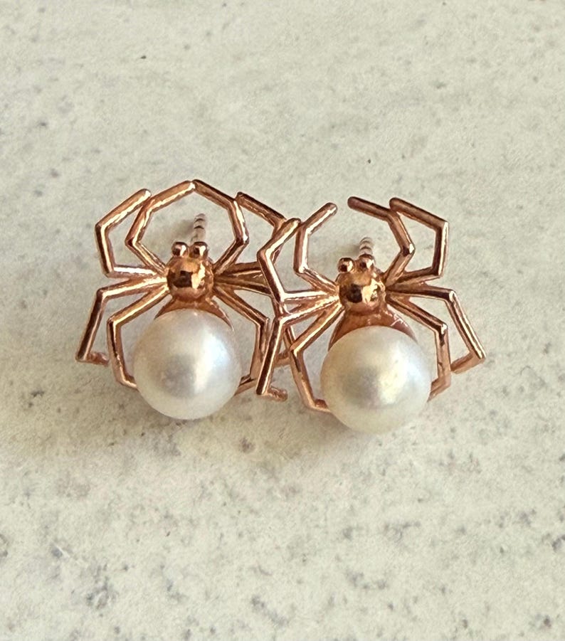 Op de afbeelding: Een paar oorknopjes in de vorm van spinnen, met poten van ros&eacute;goudkleurig metaal en een rond, wit parellichaam. Het spinnenontwerp bevat een kleine, ronde kop en acht hoekige poten die uitsteken vanaf het centrale parel-achterlijf.