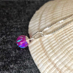 May include: A silver necklace featuring a round, iridescent, magenta-colored pendant. The pendant has a silver cap and a chain with a square link design. The necklace is displayed on a seashell against a dark background.