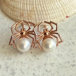 Op de afbeelding: Een paar oorknopjes in de vorm van spinnen, gemaakt van ros&eacute;goudkleurig metaal. Elke spin heeft een ronde, witte parel als achterlijf, met dunne, hoekige metalen poten die naar buiten uitsteken vanaf een centraal lichaamssegment.