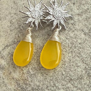 Peut inclure: Boucles d'oreilles argentées avec un motif de soleil, des pierres transparentes et des pendentifs jaunes en forme de goutte. Les boucles d'oreilles ont un détail enroulé de fil. Présentées sur une surface grise texturée.