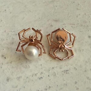 Op de afbeelding: Een paar oorknopjes in de vorm van spinnen van ros&eacute;goud. De ene oorbel heeft een ronde witte parel als achterlijf van de spin, terwijl de andere een massief ros&eacute;gouden silhouet van een spin is met zichtbare metalen poten.