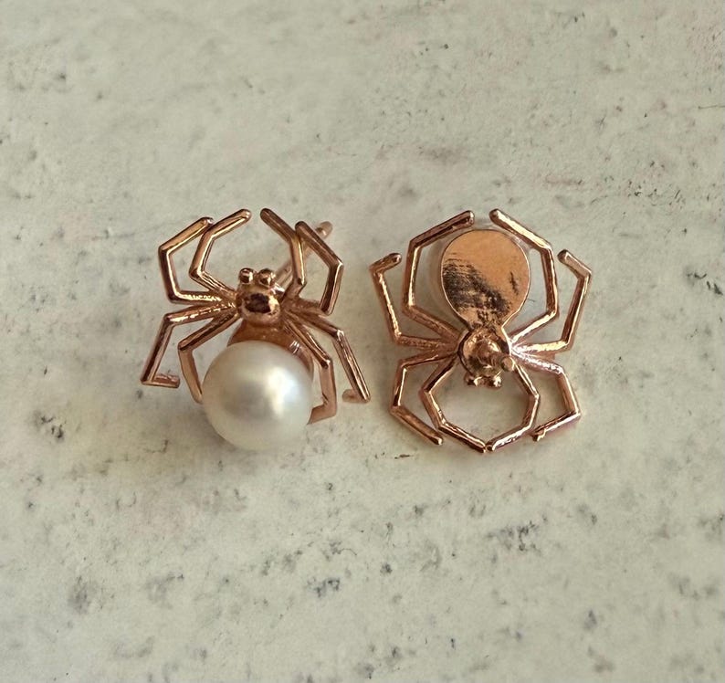 Op de afbeelding: Een paar oorknopjes in de vorm van spinnen van ros&eacute;goud. De ene oorbel heeft een ronde witte parel als achterlijf van de spin, terwijl de andere een massief ros&eacute;gouden silhouet van een spin is met zichtbare metalen poten.