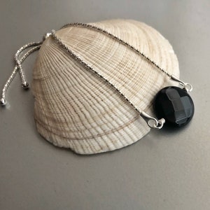 May include: A black gemstone bracelet with a silver chain and adjustable clasp. The bracelet is on a seashell background.