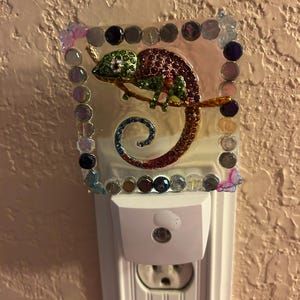 May include: A white night light with a decorative, jeweled chameleon design. The chameleon is perched on a branch and is adorned with green, red, and gold rhinestones. The night light is framed with colorful beads and gems.