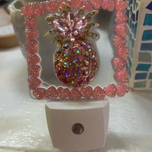 May include: A decorative night light featuring a pink pineapple design with iridescent accents. The pineapple is set within a clear square frame adorned with pink heart-shaped embellishments. The base of the night light is white.