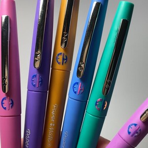May include: Six colorful pens with silver accents and a monogrammed "CH" on each. The pens are labeled "Paper Mate" and "Flair Medium".
