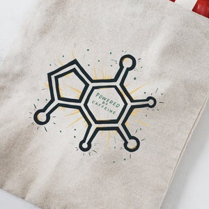 Op de afbeelding: Een beige canvas draagtas gevuld met verse producten, waaronder bananen, broccoli, een perzik, tomaten en courgette. De tas heeft een zwart-witte afbeelding van een cafeïnemolecuul met de tekst "POWERED BY CAFFEINE".