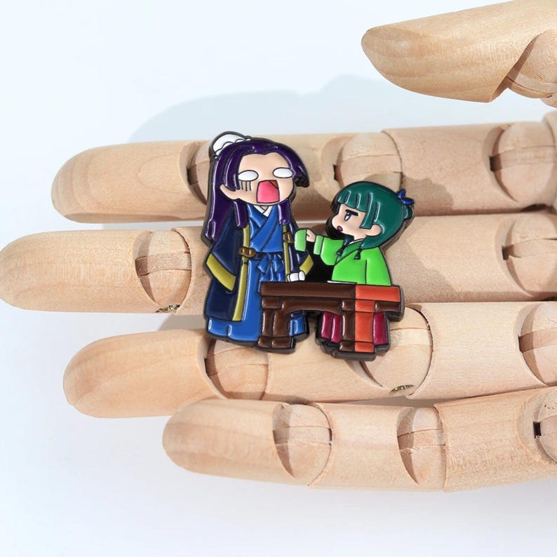 May include: A colorful enamel pin featuring two anime-style characters. One character is in blue and gold robes, the other in green. They are positioned near a small brown table. The pin is set against a wooden background.