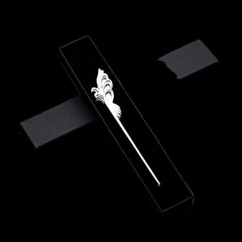May include: A black rectangular box with a white decorative hair stick. The hair stick features an ornate, stylized floral design at the top and a long, slender stem. The background is solid black.