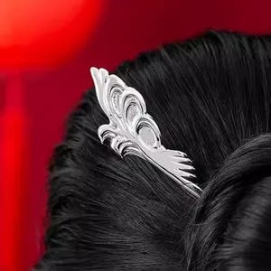 May include: A silver hair pin with an ornate, stylized feather design. The hair accessory is set against a backdrop of dark hair and a red background, creating a striking contrast. The pin is likely used for decorative purposes.