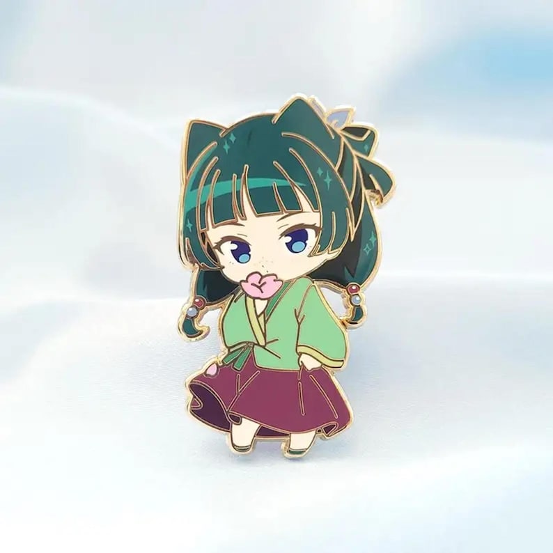 May include: A gold-colored enamel pin featuring a chibi-style anime character. The character has green hair, blue eyes, and wears a green top and a maroon skirt. The character is holding a pink flower. The pin has a gold outline.