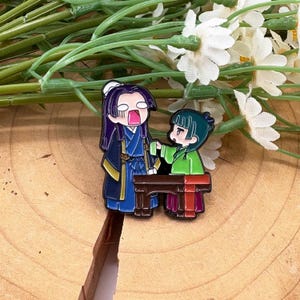 May include: Enamel pin featuring two anime-style characters. One character is in a blue and gold outfit, with purple hair and an open mouth. The other character is in a green outfit, with green hair. They are standing near a brown table.