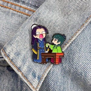 May include: A colorful enamel pin featuring two anime-style characters. One character is in a blue and yellow outfit, with purple hair and an open mouth. The other character is in a green and red outfit, pointing at the other character.
