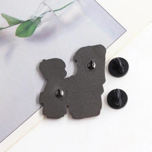 May include: A dark gray enamel pin with two figures, and two black pin backs. The pin features a design of two figures embracing. The pin is on a white and gray surface.