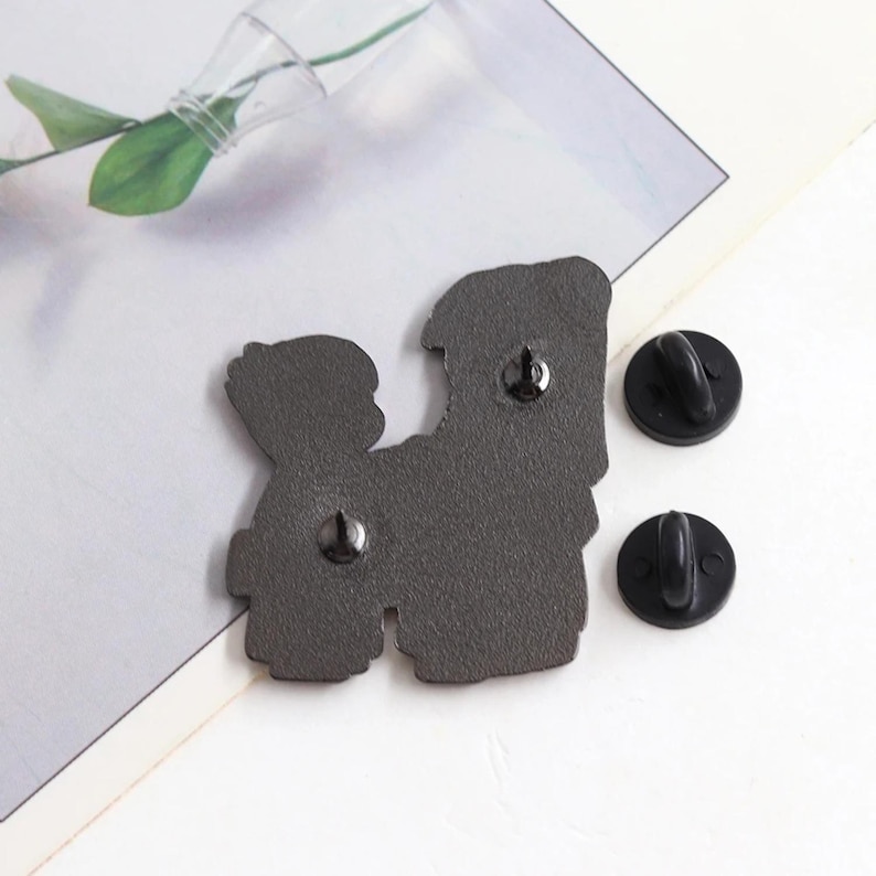 May include: A dark gray enamel pin with two figures, and two black pin backs. The pin features a design of two figures embracing. The pin is on a white and gray surface.