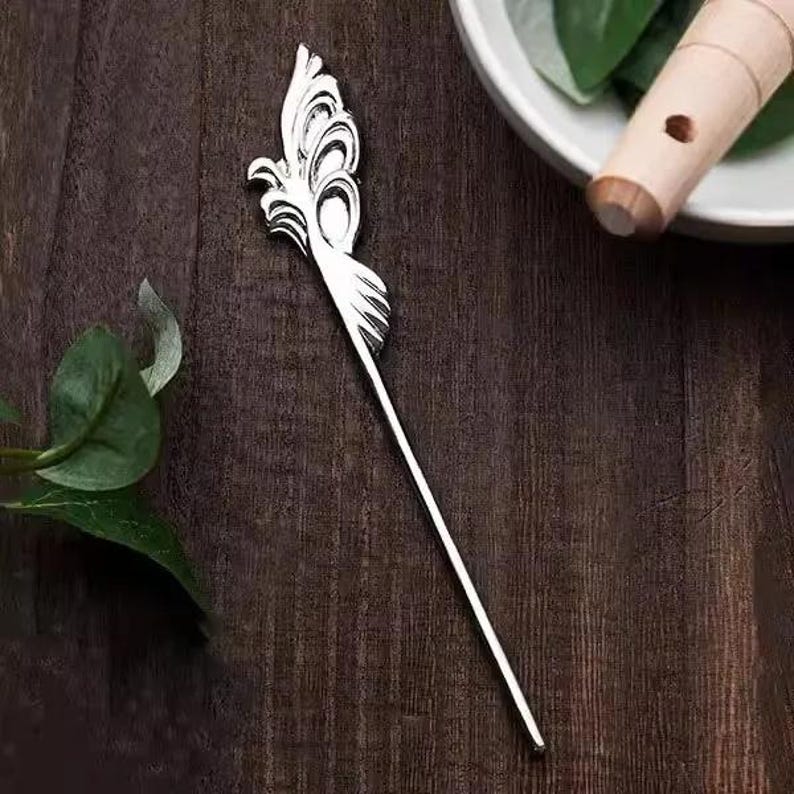 May include: A silver hair stick with a decorative feather design. The hair accessory is on a dark wood surface. The feather design has curved lines and a pointed tip. A wooden flute and green leaves are in the background.