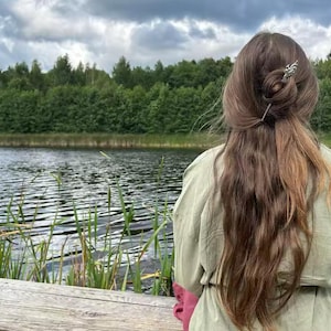May include: A person with long brown hair styled in a bun, adorned with a decorative hair pin, is seen from the back. They are wearing a green jacket with pink sleeves, and are seated on a wooden dock overlooking a lake and lush green trees.