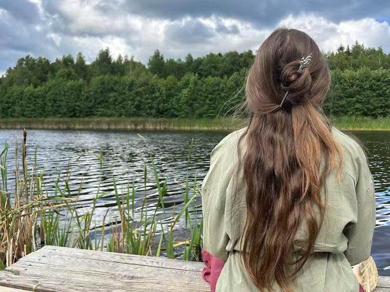 May include: A person with long brown hair styled in a bun, adorned with a decorative hair pin, is seen from the back. They are wearing a green jacket with pink sleeves, and are seated on a wooden dock overlooking a lake and lush green trees.