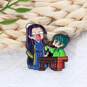 May include: Enamel pin featuring two anime-style characters. One character has purple hair and is wearing a blue and gold outfit, with an open mouth. The other character, with green hair, is holding a cup. A small brown table is between them.