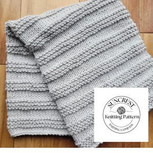 May include: A light gray knitted scarf with a ribbed design. The scarf is folded and lies on a wooden surface. A white square with a circular logo that says "Suncrest Knitting Pattern Knitting Company" is in the bottom right corner.