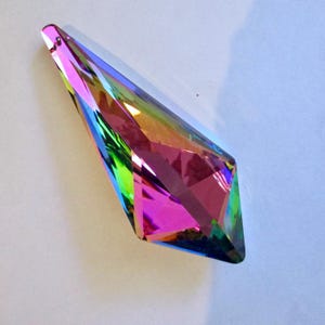 May include: A faceted, teardrop-shaped crystal pendant featuring an iridescent finish that displays a spectrum of vibrant colors including pink, green, blue, and yellow. The glass object has a small hole at the top for hanging or attachment.