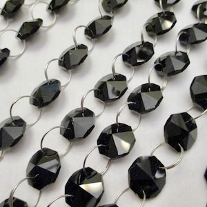 May include: Close-up of multiple strands of black faceted beads connected by silver-colored metal rings. The beads have a glossy finish and are arranged in parallel rows, creating a repeating pattern. The background is a light, neutral color.