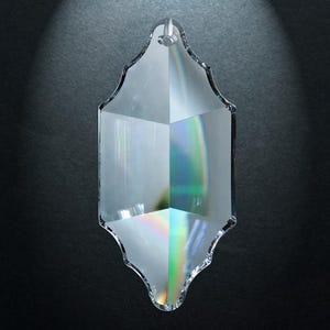 May include: A clear, faceted crystal pendant with a pointed, elongated shape and scalloped edges. The glass features a small circular hole at the top for hanging and displays subtle prismatic light refractions across its smooth, polished surface.
