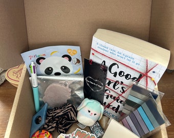 Puede incluir: Una caja de cartón llena de varios artículos, incluyendo un libro titulado "A Good Girl's Guide to Murder", una mascarilla facial de panda, un bolígrafo multicolor, un conejito de peluche, pegatinas y un bálsamo labial. La caja también contiene un marcapáginas y una goma para el pelo.