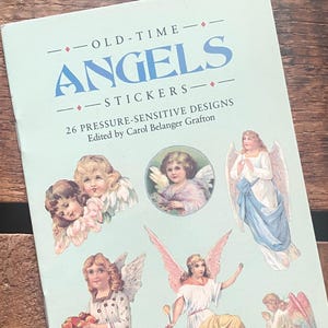 Puede incluir: Un libro de pegatinas azul claro titulado "OLD-TIME ANGELS STICKERS" con ilustraciones de ángeles. El libro contiene 26 diseños sensibles a la presión, editados por Carol Belanger Grafton. Está sobre una superficie de madera.
