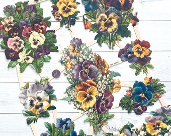 Puede incluir: Surtido de flores de papel vintage con pétalos morados, amarillos y blancos. Las flores están dispuestas en un patrón circular sobre un fondo de madera blanco.