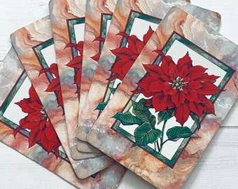 Könnte beinhalten: Ein Stapel rechteckiger Untersetzer mit einem Weihnachtsstern-Blumenmuster. Jeder Untersetzer zeigt einen leuchtend roten Weihnachtsstern, der von einem grünen Rahmen umgeben ist, vor einem marmorierten Hintergrund mit Orange-, Grau- und Weißtönen. Die Untersetzer liegen auf einer weißen Holzoberfläche.